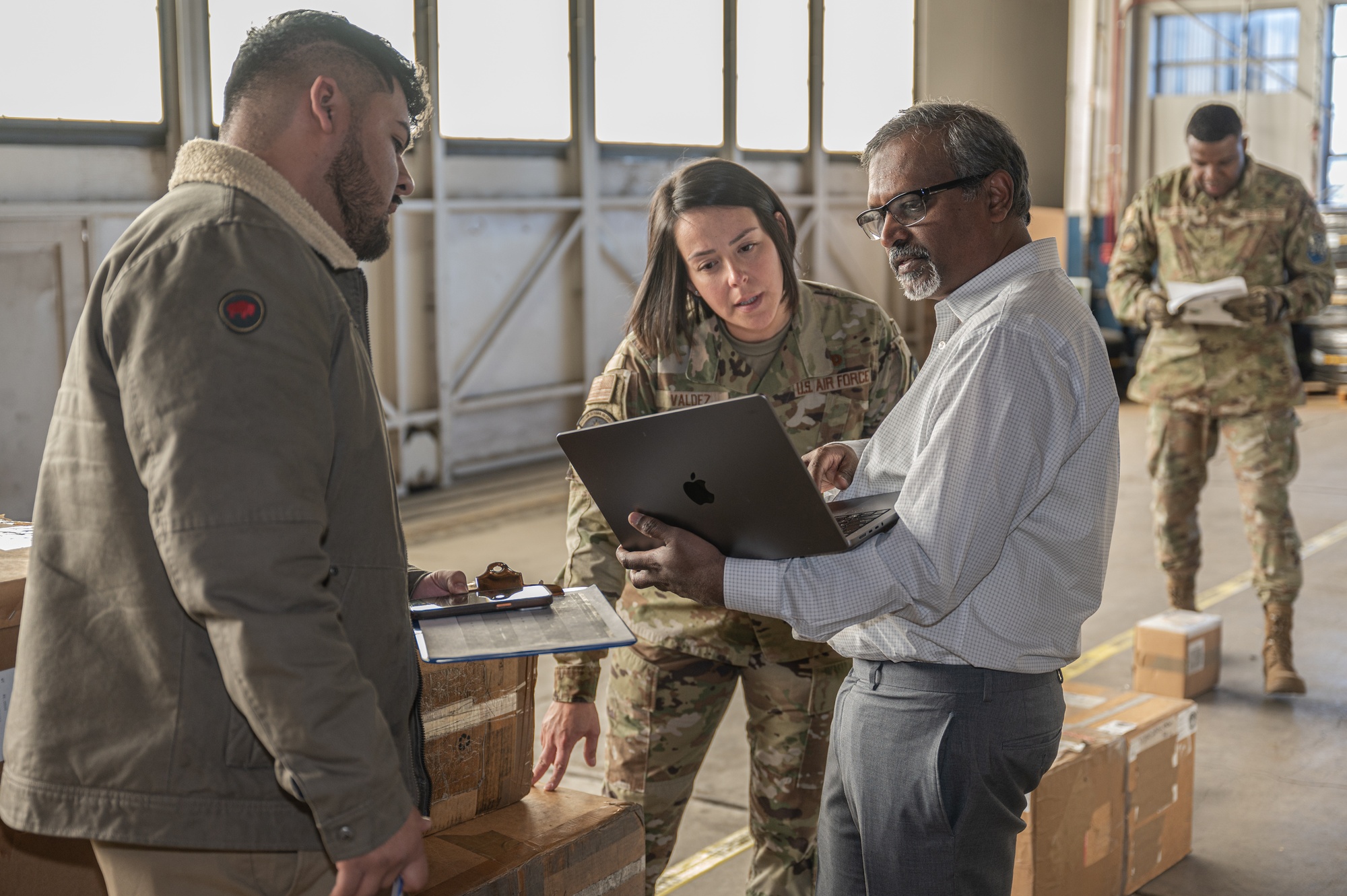 An Air Force logistics specialist and two civilian colleagues review a laptop beside stacked cargo boxes.