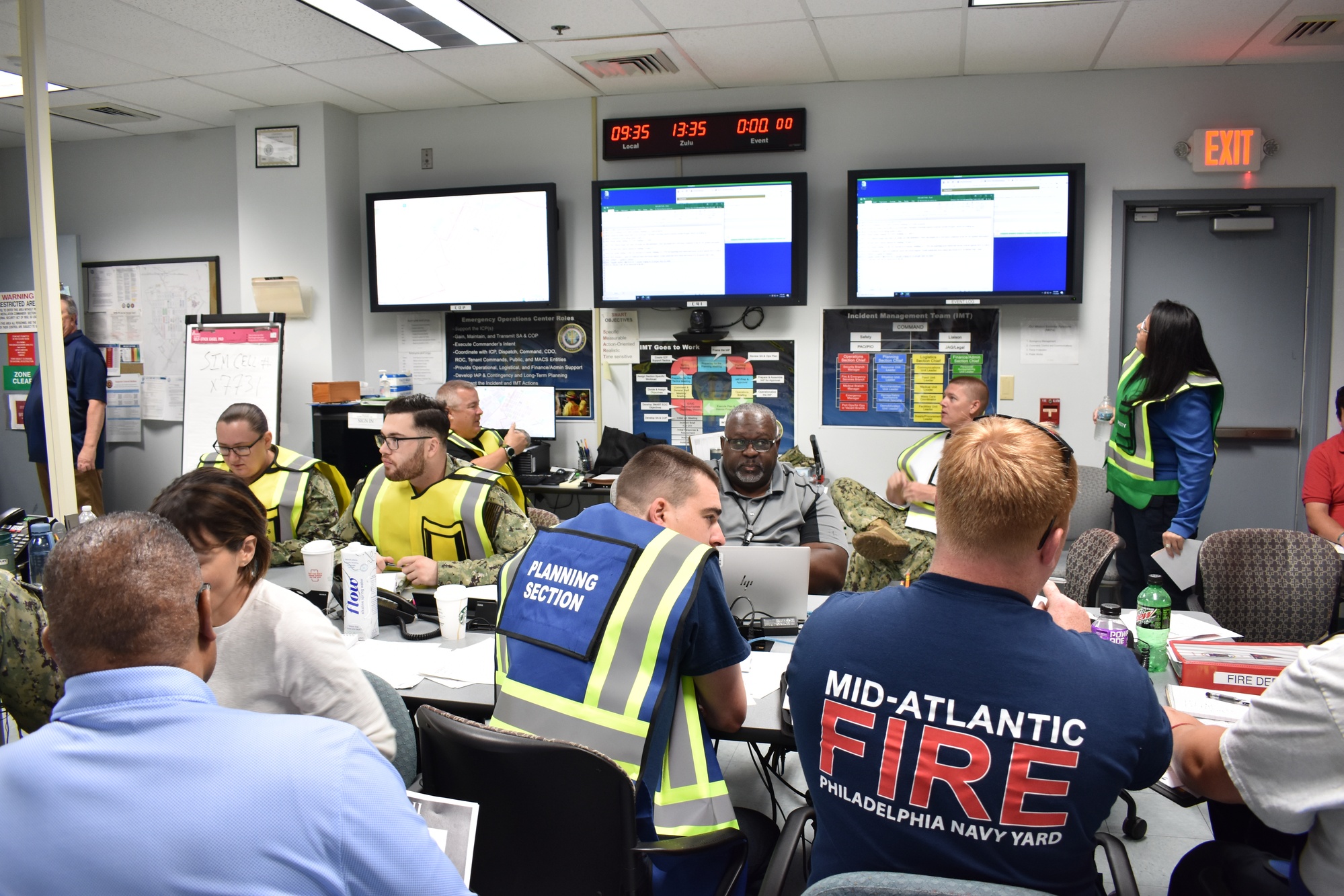 Emergency operations personnel coordinate around tables and laptops beneath wall-mounted status screens.