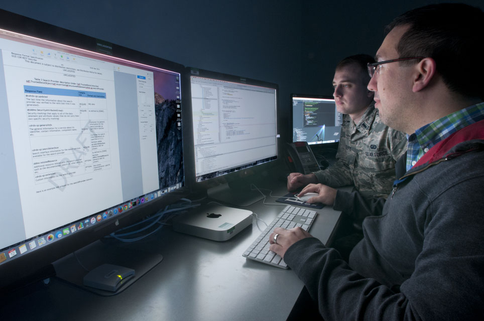A uniformed test engineer and a civilian developer review integration results across several computer monitors.