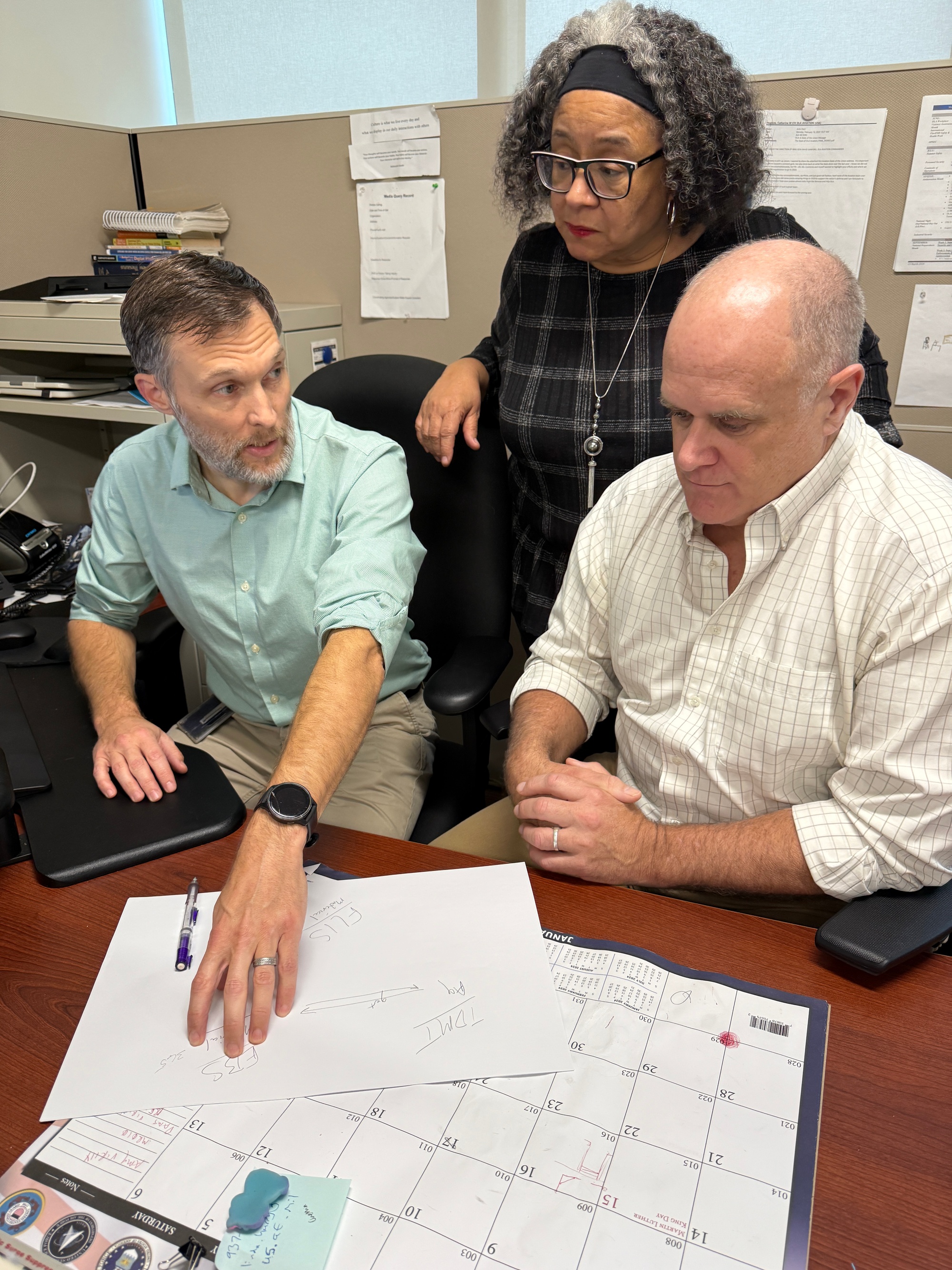 Three Defense Logistics Agency technology colleagues review a hand-drawn systems diagram and project calendar.