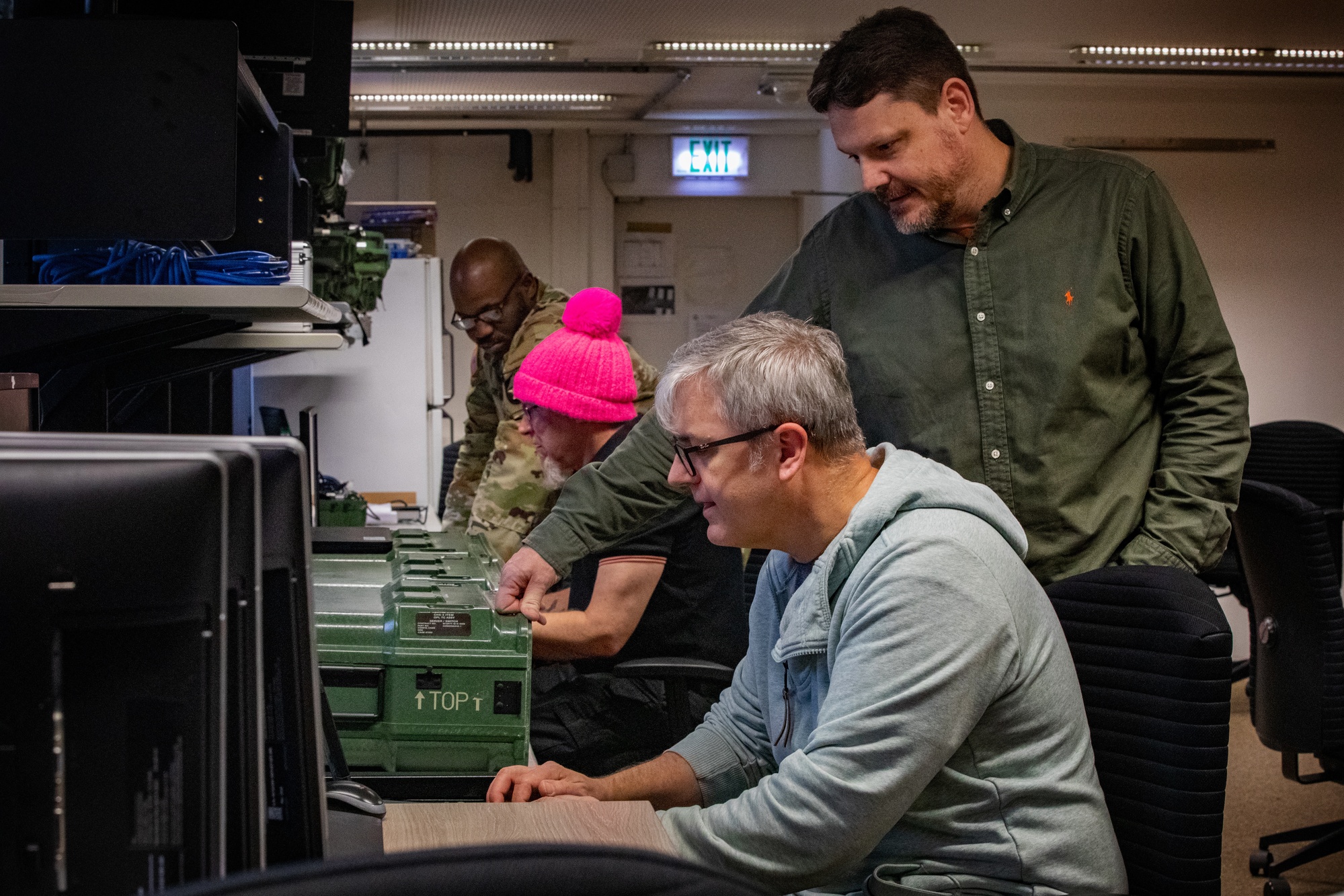 Civilian developers and a uniformed soldier work together at computers and equipment in a technical workshop.